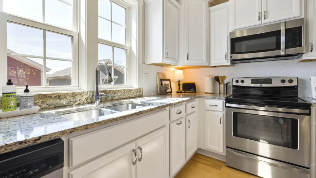 
Alt Text: Sunlit kitchen corner showcasing smoothly repainted kitchen cupboards in bright white, paired with granite countertops and a stainless steel double sink
