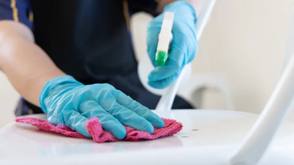 Gloved hands wiping down a white surface with a cloth and disinfectant spray