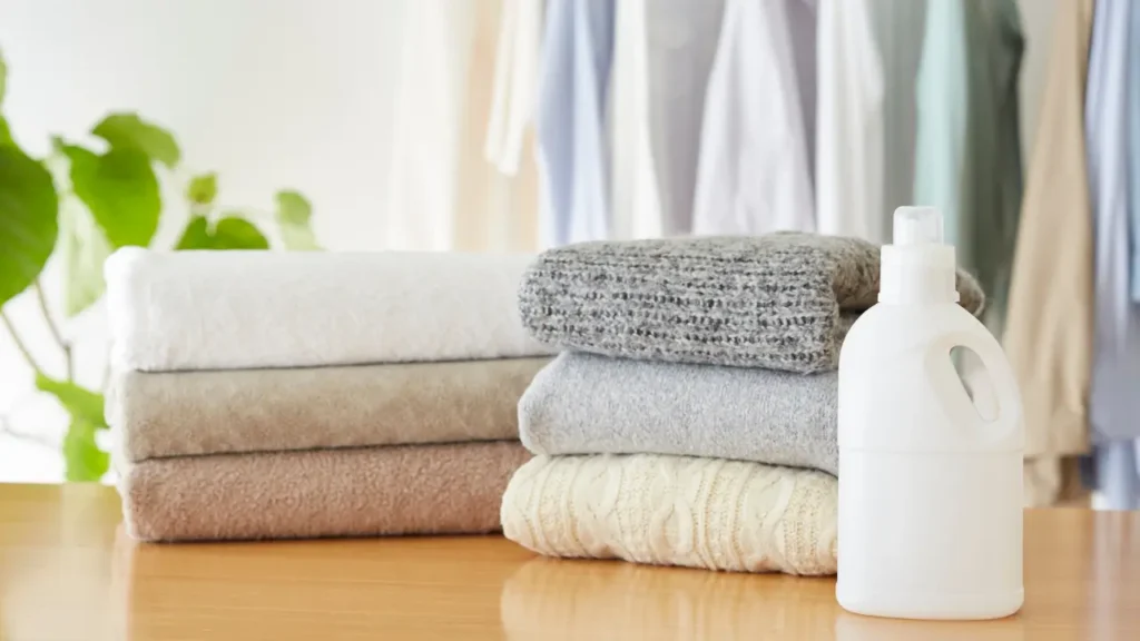 Neatly folded towels and sweaters stacked on a wooden table next to a white detergent bottle