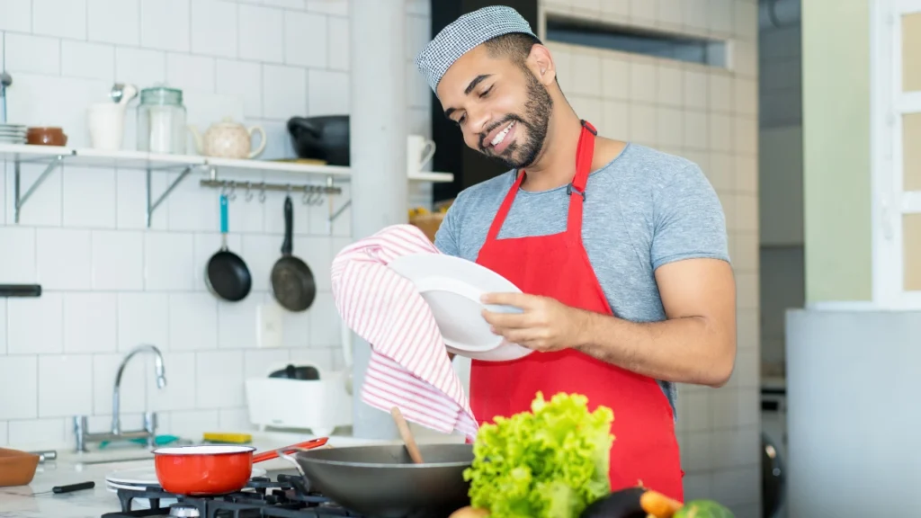 Smiling man in a red apron drying a plate in a kitchen while cooking