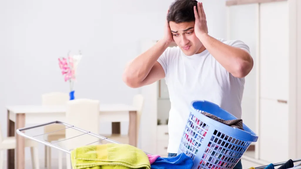 Frustrated man holding his head while looking at an overflowing blue laundry basket next to a drying rack