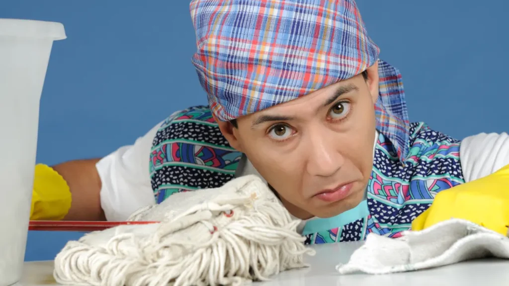 Man in a plaid bandana leaning on a table next to a mop and cleaning rag, looking tired