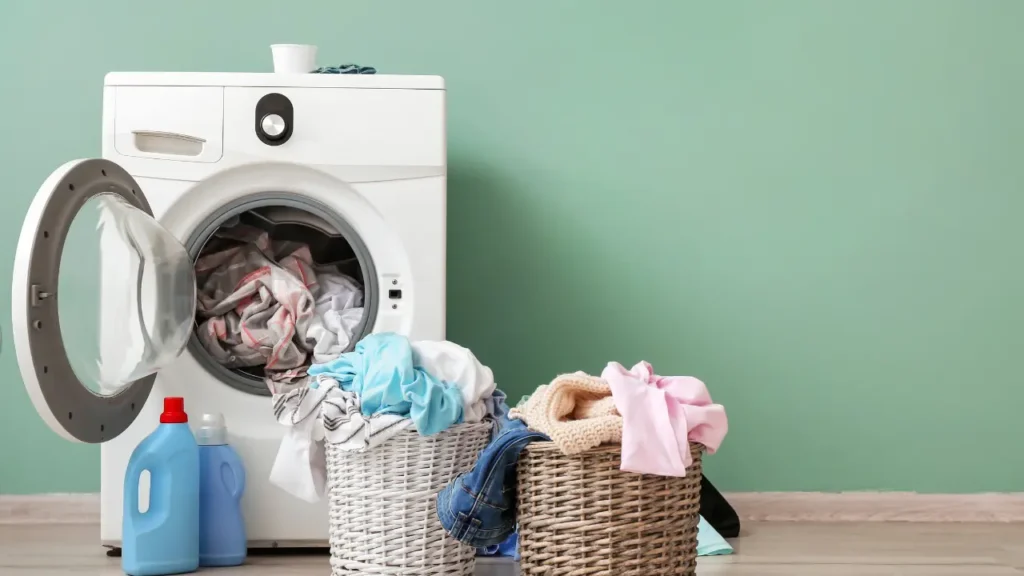 Open washing machine stuffed with clothes next to detergent bottles and two wicker laundry baskets against a green wall