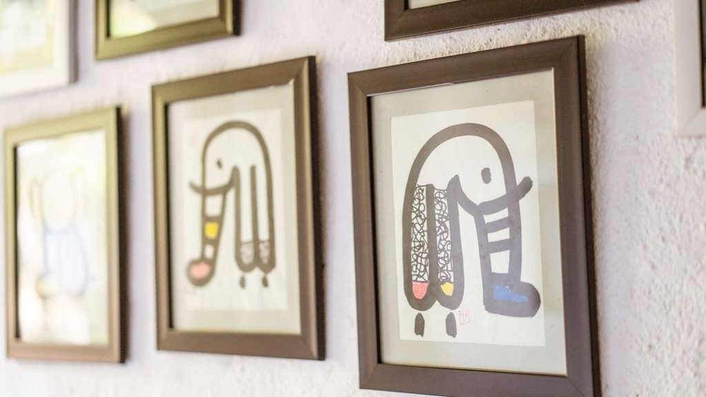 Close-up of framed children's drawings displayed in a row of brown and gold frames on a textured white wall