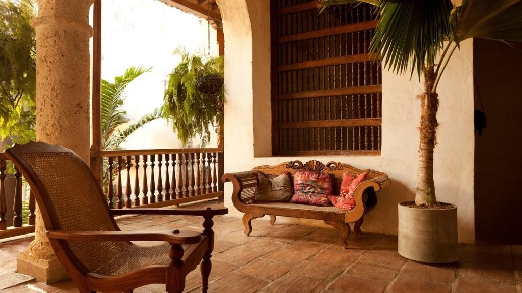 Colonial-style veranda with a carved wooden rattan bench, cane armchair, and terracotta tile flooring beside a potted palm