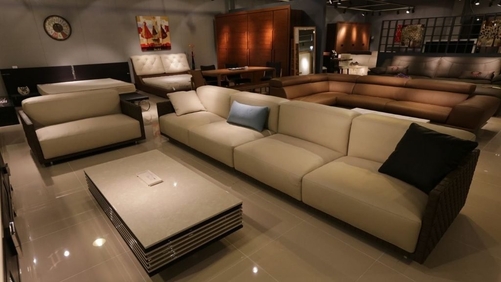Furniture showroom floor displaying a cream leather sectional sofa, glass coffee table, and brown leather sofas in the background
