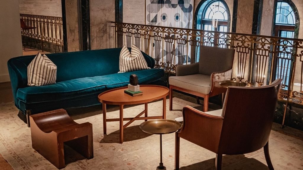 Boutique hotel lounge with a teal velvet sofa, striped throw pillows, leather armchairs, and an ornate brass balcony railing