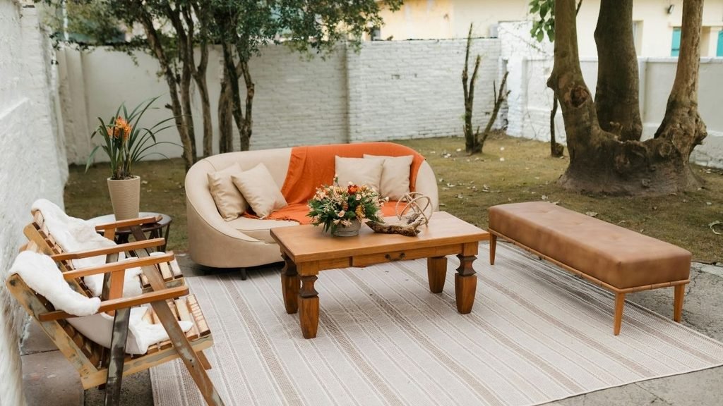 Outdoor patio lounge area with a curved beige sofa, orange throw blanket, wooden coffee table, and leather bench on a striped rug