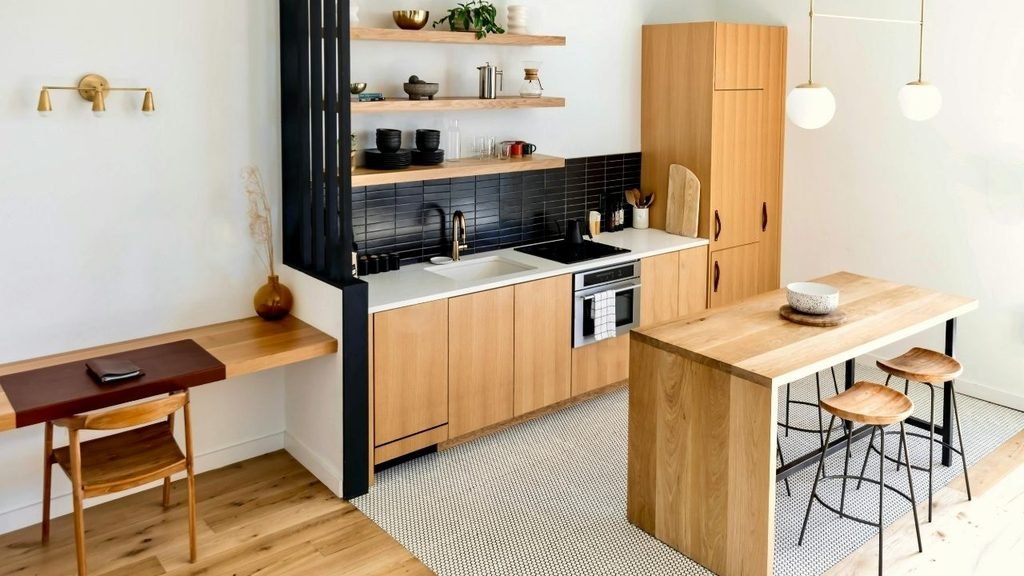 Compact modern kitchen with light oak cabinetry, black tile backsplash, open shelving, and a wooden breakfast bar with stools