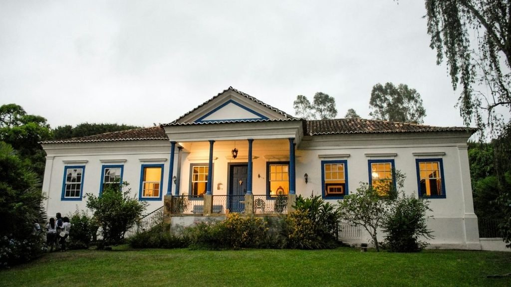 White colonial farmhouse with blue window trim, a columned front porch, terracotta roof, and green lawn at dusk