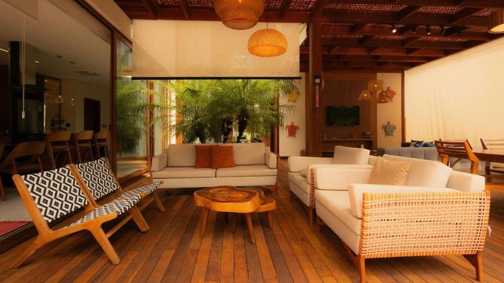 Warm wood-toned living room with cream sofas, woven rattan pendant lights, and a view of indoor palm trees through glass doors