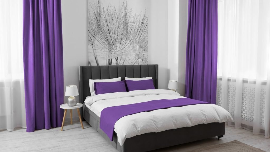 Bedroom styled with a connected purple color palette across floor-length curtains, bedding, and pillows against a gray tufted headboard