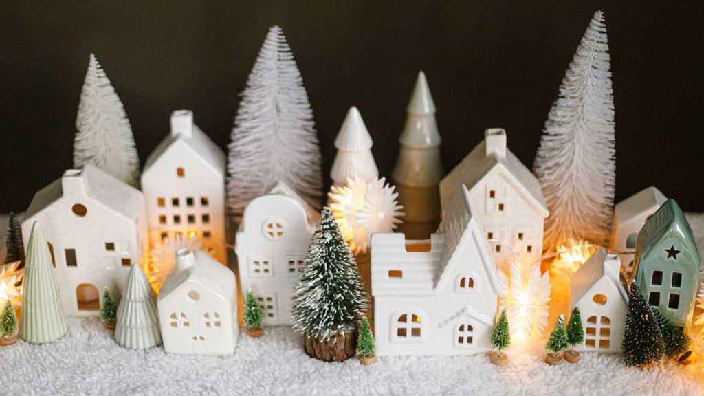 Small white ceramic house village lit from within by warm string lights, surrounded by miniature bottle-brush trees