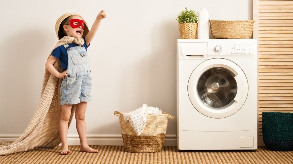 Little girl in a superhero cape and mask playing next to a front-load washing machine in a laundry room
