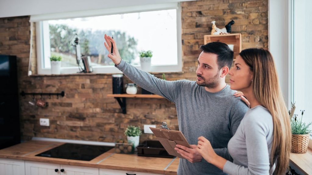 A man pointing out details while a woman holds a clipboard, standing together in a rustic modern kitchen during a home inspection or renovation planning.