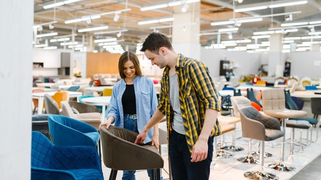 Couple shopping for bedroom furniture in a home furnishing store, comparing velvet chairs and modern deco