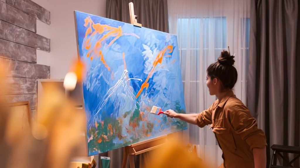 A female artist with her hair in a bun painting a vibrant blue and orange abstract artwork on a large canvas mounted on a wooden easel in a studio
