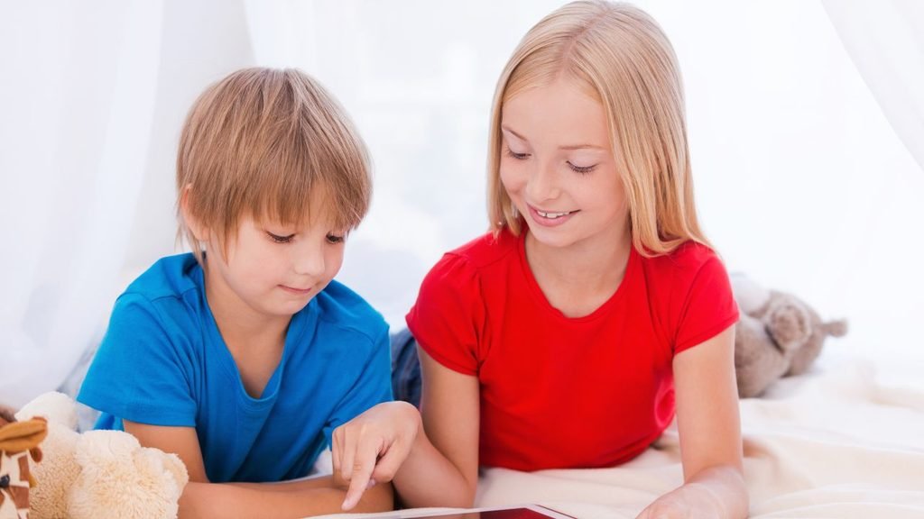 Two children lying on a cozy bed under a white canopy, looking at a tablet with plush toys nearby