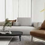 Living room with sheer curtains, a glass vase on a black coffee table, and a tan leather accent chair beside a slim gold floor lamp