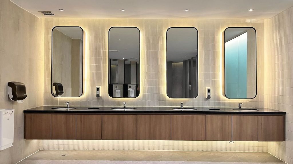 A clean, modern commercial restroom featuring a long wooden vanity, black countertop, four sinks, and tall rectangular mirrors with warm backlighting.