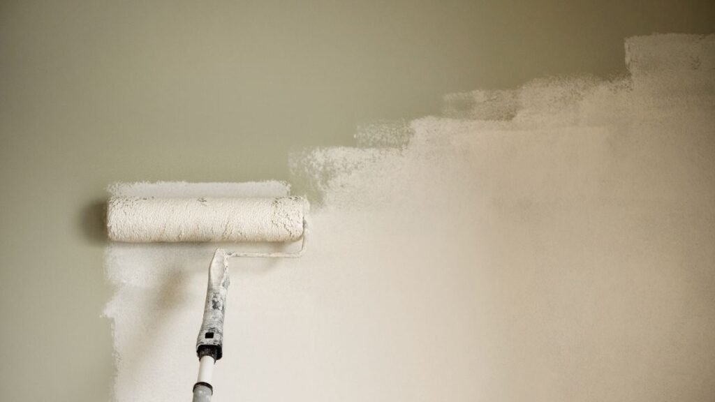 A close-up of a paint roller applying a fresh coat of white paint over a beige wall.