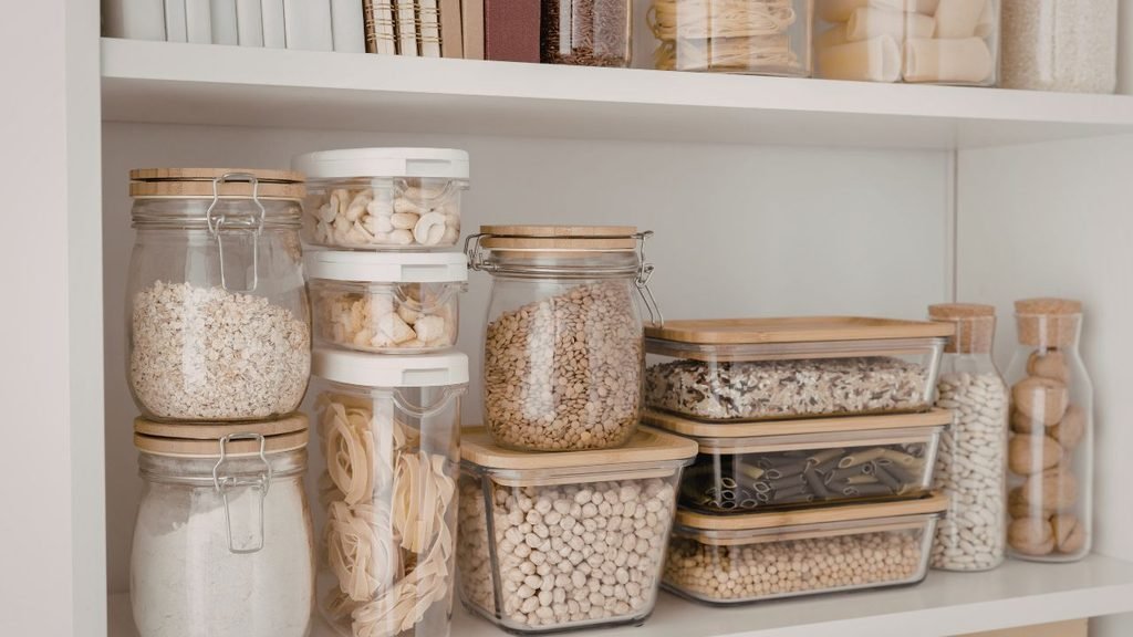 Organized pantry shelf with glass jars and bamboo-lid containers filled with pasta, grains, and beans