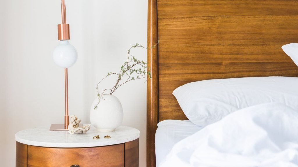 Round marble-top nightstand with a copper table lamp and white vase beside a walnut wood headboard