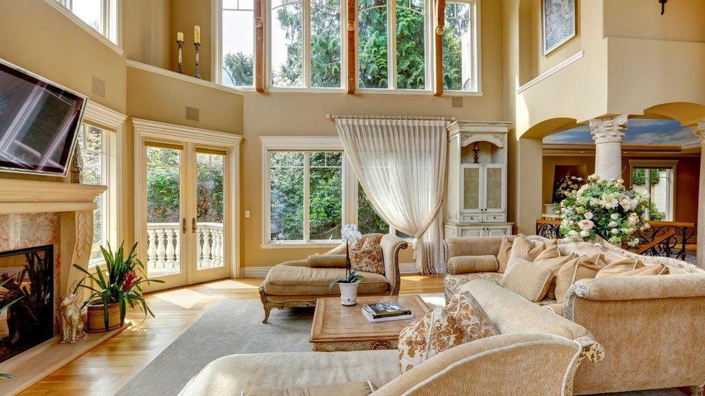 Bright two-story living room with stone fireplace, floor-to-ceiling windows, cream upholstered furniture, and fresh flower centerpiece