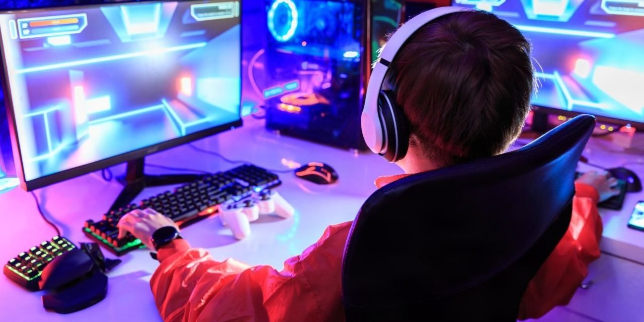 Gamer wearing white headphones at a dual-monitor RGB PC in a small room gaming setup with a gamepad and keypad