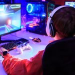 Gamer wearing white headphones at a dual-monitor RGB PC in a small room gaming setup with a gamepad and keypad