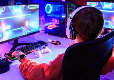 Gamer wearing white headphones at a dual-monitor RGB PC in a small room gaming setup with a gamepad and keypad