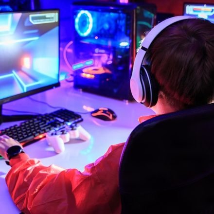 Gamer wearing white headphones at a dual-monitor RGB PC in a small room gaming setup with a gamepad and keypad