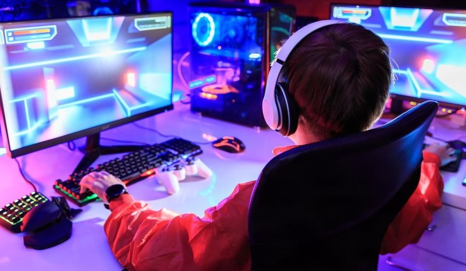 Gamer wearing white headphones at a dual-monitor RGB PC in a small room gaming setup with a gamepad and keypad