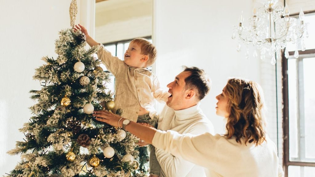 Family of three decorating a frosted Christmas tree with gold and white ornaments while the toddler reaches for the tree topper