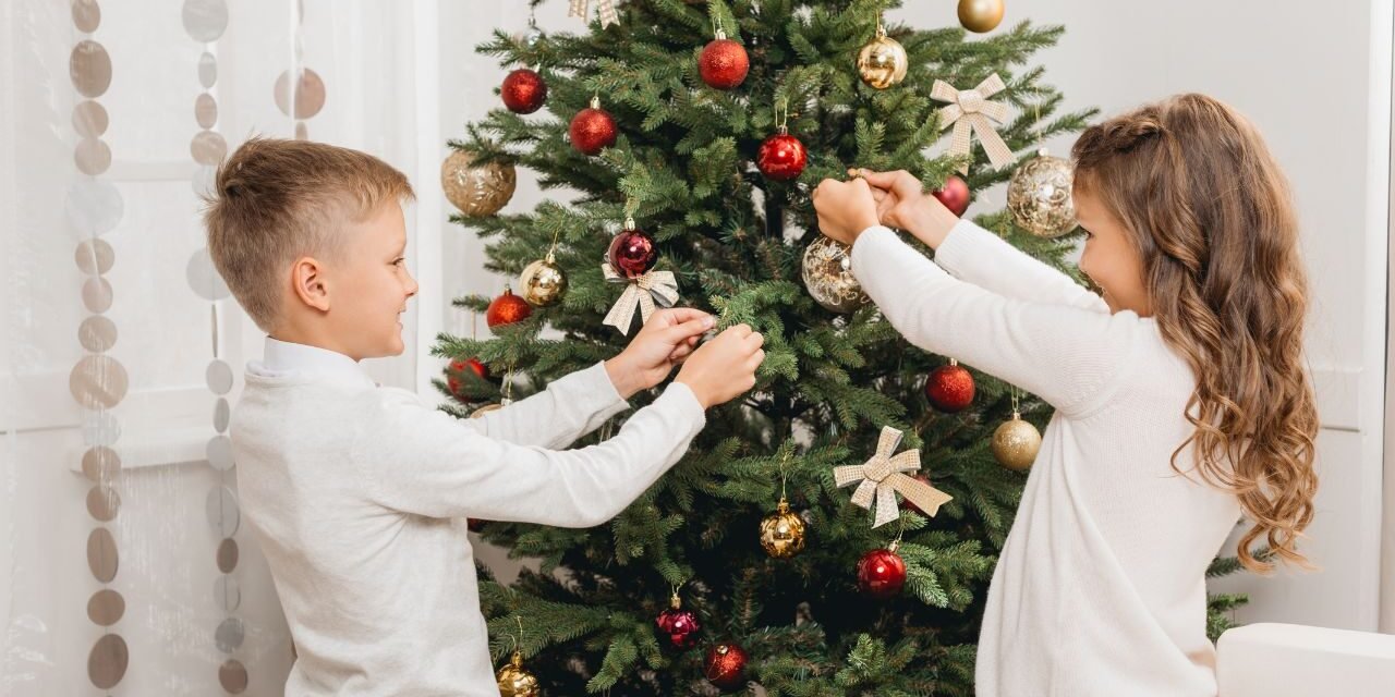 Boy and girl decorating a green Christmas tree with red and gold baubles and ribbon bows, a fun christmas tree design idea for kids
