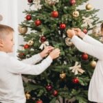 Boy and girl decorating a green Christmas tree with red and gold baubles and ribbon bows, a fun christmas tree design idea for kids