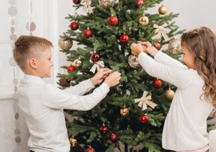 Boy and girl decorating a green Christmas tree with red and gold baubles and ribbon bows, a fun christmas tree design idea for kids