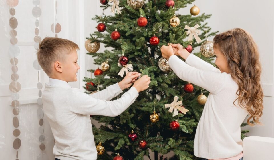 Boy and girl decorating a green Christmas tree with red and gold baubles and ribbon bows, a fun christmas tree design idea for kids