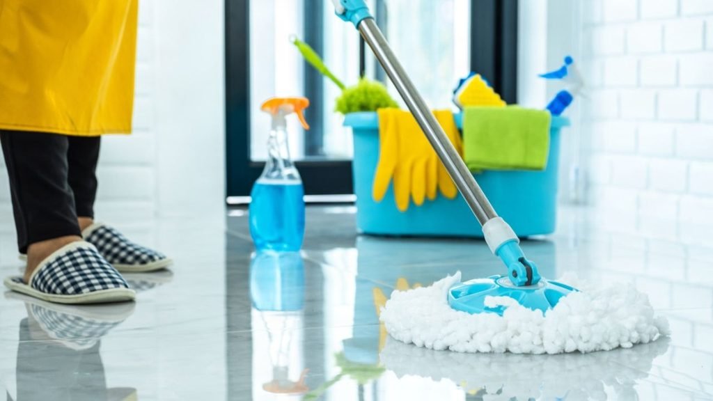 Spin mop on a glossy tiled floor beside a spray bottle and a bucket of gloves and cloths, ideal for apartment floor cleaning