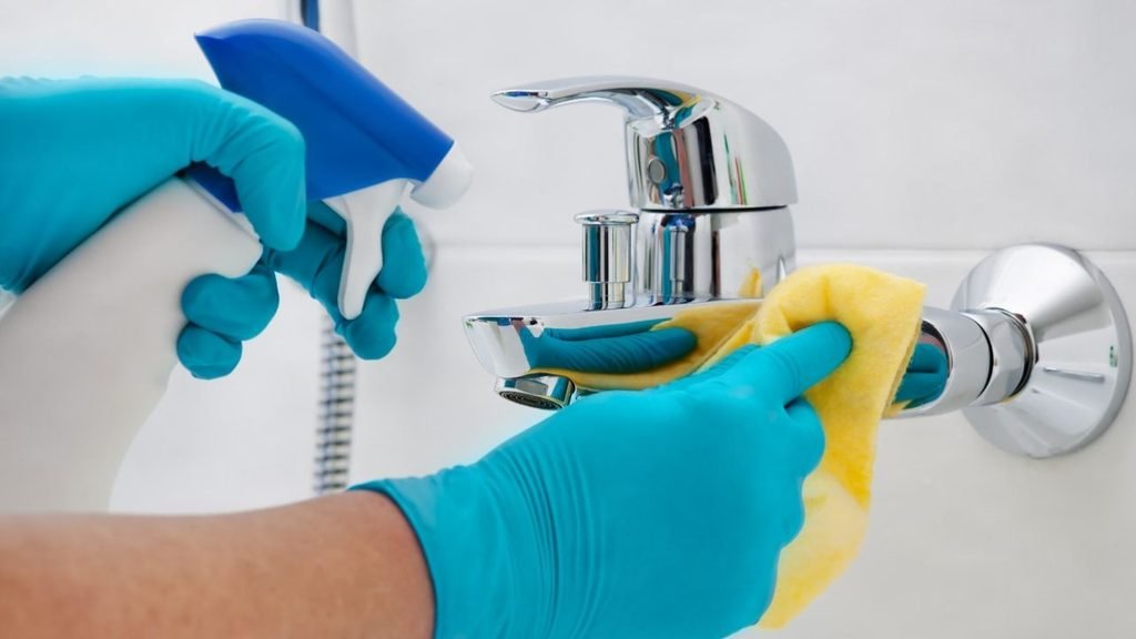 Hands in teal gloves polishing a chrome bathroom faucet with a yellow cloth and spray cleaner