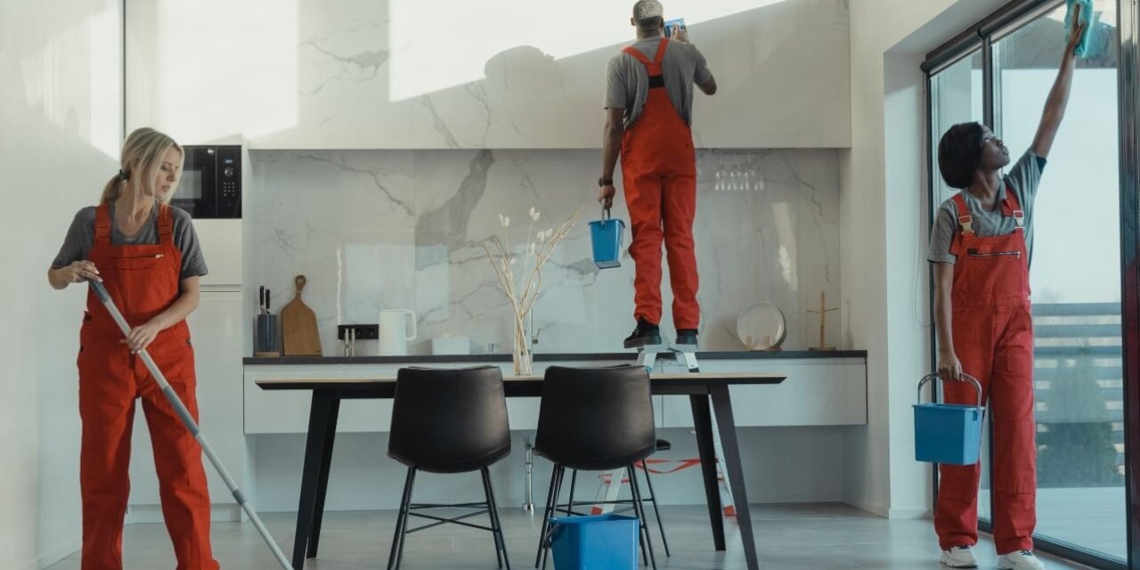Three cleaners in red overalls mopping, wiping cabinets and washing windows during a deep cleaning of a modern kitchen