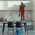 Three cleaners in red overalls mopping, wiping cabinets and washing windows during a deep cleaning of a modern kitchen