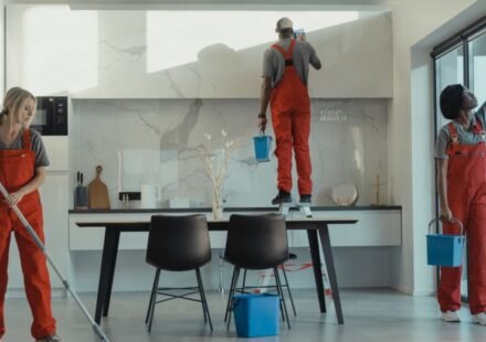Three cleaners in red overalls mopping, wiping cabinets and washing windows during a deep cleaning of a modern kitchen