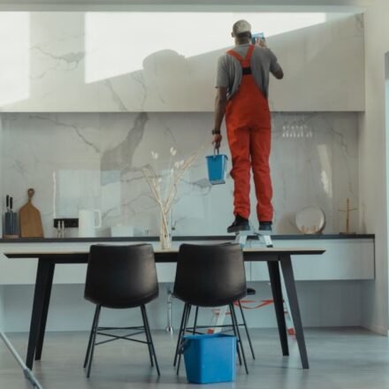 Three cleaners in red overalls mopping, wiping cabinets and washing windows during a deep cleaning of a modern kitchen