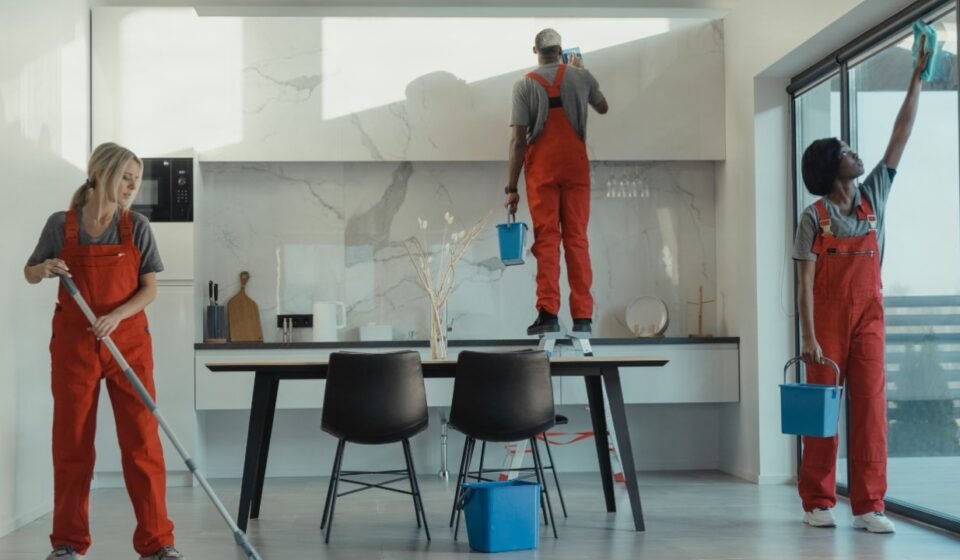 Three cleaners in red overalls mopping, wiping cabinets and washing windows during a deep cleaning of a modern kitchen