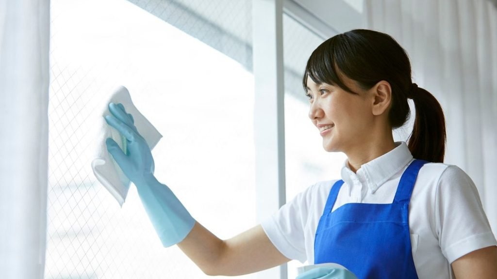 Smiling woman in a blue apron and gloves wiping a window with a white cloth, a daily cleaning habit for the home