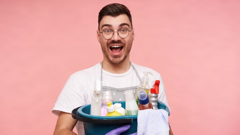 Excited man in glasses holding a bucket of cleaning sprays, cloths and gloves, ready to try new cleaning hacks and tips