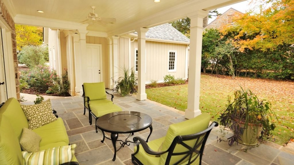 Covered stone patio with lime green cushioned seating and a round fire pit table next to a lawn covered in fall leaves