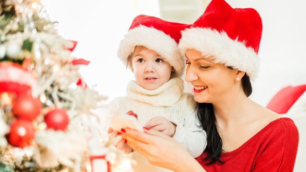 Mother and toddler in Santa hats hanging a handmade ornament on a white flocked tree, a cozy DIY christmas tree design idea