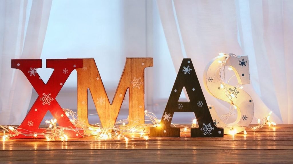 Colorful wooden XMAS letters with snowflake designs and warm glowing string lights on a wooden surface in front of white curtains